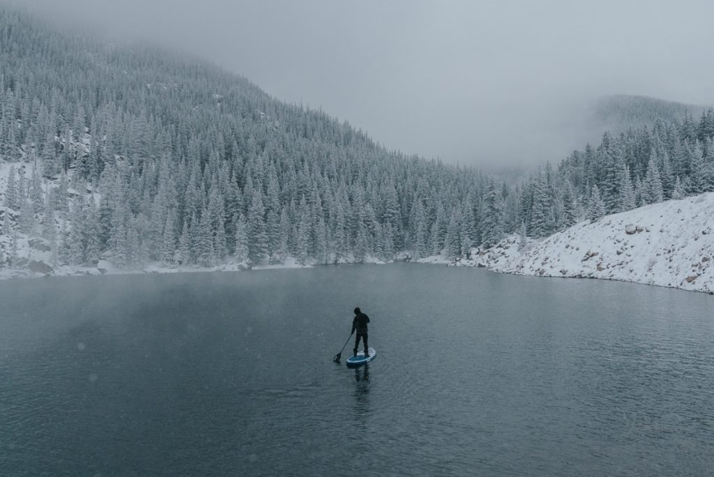 Paddle Boarder in the Middle of a Wintry Scene with Water, Trees, Mountains, and Snow