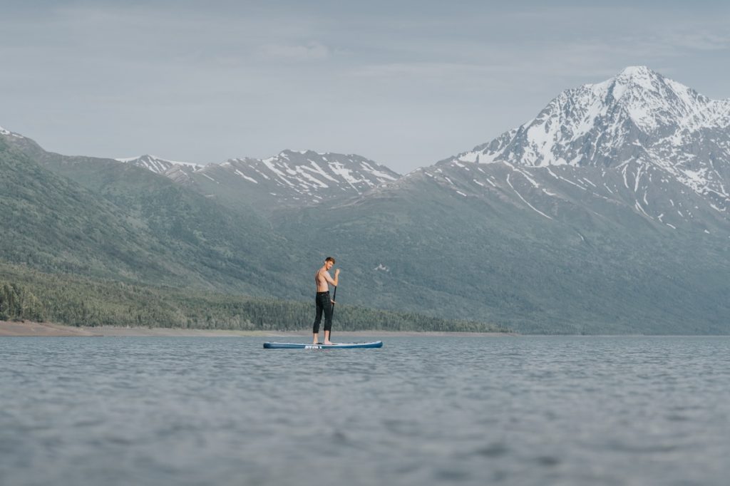Paddle Boarder Holding Atoll Board in Front of Water and Mountains