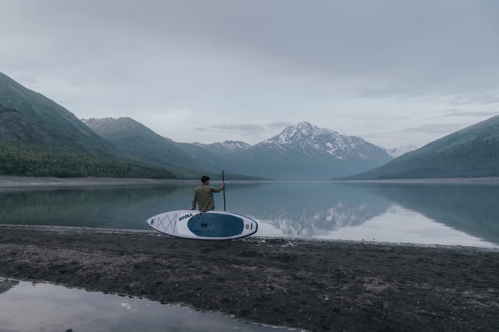 Rear View of Paddler Boarder Resting on Atoll Board in Front of Mountain and Water